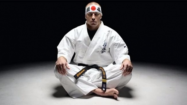 Крупный план Жоржа Сен-Пьера (Georges St-Pierre), сидящего в традиционной позе сэйдза на полу на белом студийном фоне. На нем белоснежное плотное доги (кимоно) для каратэ Киокушинкай с аутентичной иероглифической вышивкой «Кёкусин» синими нитями на левой стороне груди. На талии повязан потертый черный пояс (дан), завязанный правильным узлом. На голове бойца — белая повязка хатимаки с изображением красного круга (японского флага Хиномару) по центру. Руки Сен-Пьера сжаты в кулаки и спокойно лежат на бедрах, взгляд сосредоточен, выражает глубокую ментальную концентрацию, дисциплину и спокойствие перед поединком.