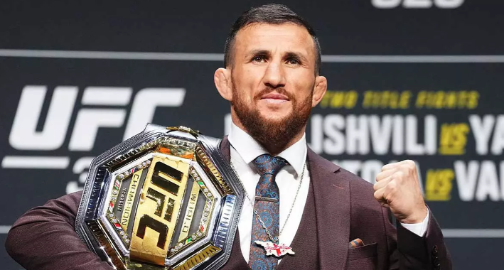A medium shot of an athletic Georgian MMA fighter with a beard, wearing a sharp brown formal suit, white shirt, and a blue patterned tie with a silver chain necklace. He is proudly holding a heavy gold and silver UFC championship belt over his right shoulder and making a fist with his left hand. Smiling confidently at a press conference. Background features a dark gray branded wall with blurry text "UFC".